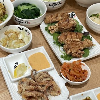 Kajiken - Fried squid and chicken wings - The image displays multiple Japanese dishes served in white bowls and plates on a wooden table. There are fried chicken pieces on a rectangular plate, accompanied by lemon and dipping sauce. Several bowls contain vegetables, kimchi, and other side dishes. The dishes are presented with Japanese-style bowls featuring black and red text. The setting suggests a communal meal with a focus on fried and pickled foods. - Ramen Near Me - Champaign, Illinois