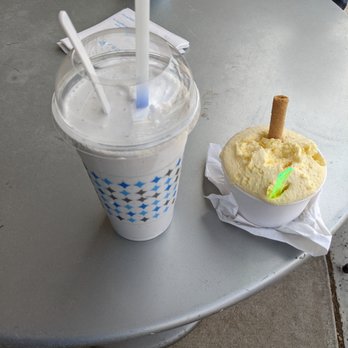 Frost Gelato - L-R: Grande Size Shake + Medium size Limoncello - A milkshake in a clear plastic cup with a domed lid and two straws, one white and one blue, is placed on a gray table. Next to it is a bowl of lemon sorbet topped with a wafer stick, resting on a napkin. In the background, there is a small cup with a blue star pattern and a book with a yellow cover featuring a pattern of red and white shapes. The setting appears to be outdoors or in a casual dining area. - Desserts Near Me - Albuquerque, New Mexico