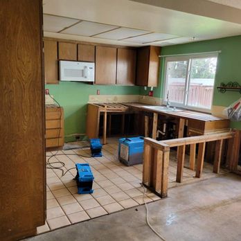 Rupprecht Construction - 2. This is what Phil started with, after the remediation team left. - The image shows a kitchen with green painted walls and wooden upper cabinets, some of which are missing doors. The countertop is partially removed, exposing the framing and plumbing underneath. There are two blue construction tools on the tiled floor, and a microwave is mounted on the wall. A sliding glass door is visible on the right, leading outside, with a plastic bag hanging on a wall-mounted shelf. The space appears to be in the process of remodeling or renovation. - General Contractors Near Me