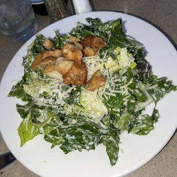 M Restaurant - Caesar salad - A Caesar salad with chopped romaine lettuce, shredded cheese, and croutons is presented on a white round plate. The salad appears to be dressed with Caesar dressing, and the croutons are scattered on top. The plate is placed on a gray surface, with glasses of water visible in the background. - Italian Near Me - Avalon, California