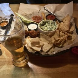 Cold Combo appetizer and a stale coors light - A tray of Mexican appetizers features tortilla chips, guacamole, salsa, and chicken meatballs, served with small bowls of dips. A glass of beer is placed in front of the tray on a wooden table, with a person partially visible in the background. The presentation includes paper lining and various condiments.