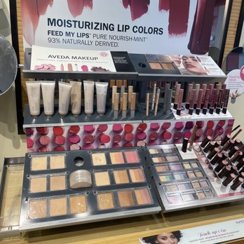 Eclipse - The image shows a makeup display on a wooden counter, featuring various cosmetic products including moisturizing lip colors, lip glosses, and eyeshadow palettes. The display includes tubes of lip gloss or liquid lipstick, small containers of lip balm or gloss, and multiple eyeshadow palettes with different shades. Promotional signage above highlights moisturizing lip colors and natural ingredients. The display is organized with products neatly arranged in rows, with a large poster in the background advertising lip colors. - Nail Salons Near Me - Tarrytown, New York