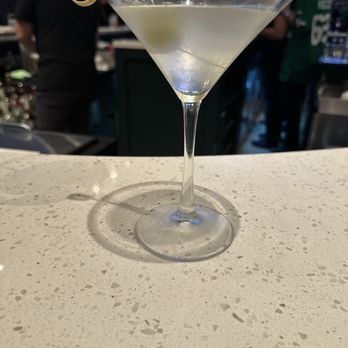 Roosters - Glenside Dirty Martini - A clear cocktail served in a martini glass with an olive garnish, placed on a bar counter with a busy bar scene in the background. - Chicken Wings Near Me - Glenside, Pennsylvania