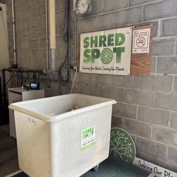 Shred Spot - Our paper getting weighed before shredding. - A large white plastic bin with a Shred Spot sticker and handwritten weight markings is positioned on a metal platform scale in an industrial environment. The background features a concrete block wall with a clock, speaker, and various electrical wires. The setting appears to be a document shredding or recycling facility, emphasizing secure disposal and environmental conservation. - Recycling Center Near Me - Northbrook, Illinois