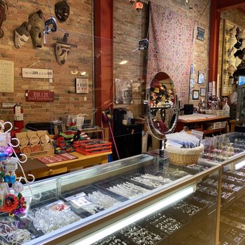 Dimple's - A jewelry display case filled with rings, bracelets, and necklaces is situated inside a gift shop with brick walls. The background features decorative masks, framed pictures, and signs, along with shelves holding various items. The shop has a warm, eclectic decor with a mix of cultural artifacts and handcrafted accessories. - Women's Clothing Near Me - Racine, Wisconsin