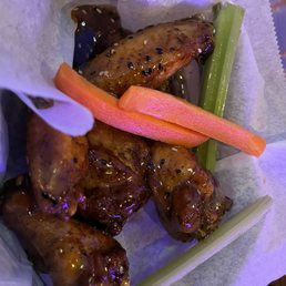 Sesame Citrus Smoked Wings - Chicken wings glazed with sesame citrus sauce, served with celery and carrot sticks on parchment paper, likely at a restaurant or bar.