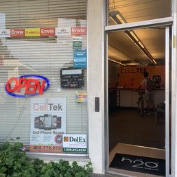 Front of store view, close to habit burger on Hollister. - The image depicts the storefront of Celltek, a mobile device repair business in Goleta, CA. The window displays an illuminated red and blue 'OPEN' sign, business hours, and various signs including advertisements for cell phone repair and financial services. The entrance is open, revealing the interior with a customer and staff member, and a mat with the 'h2o Wireless' logo is placed at the door. There are plants outside the window, and the overall scene suggests a small, local electronics repair shop.