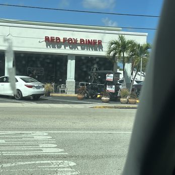 Red Fox Diner - Outside - The image depicts the exterior of Red Fox Diner, a restaurant with a white facade and red signage. There are outdoor tables and chairs, palm trees, and several parked vehicles, including a white car and a golf cart with signage. The scene is set on a sunny day with a blue sky and some clouds, and power lines are visible overhead. - Breakfast & Brunch Near Me - Lighthouse Point, Florida