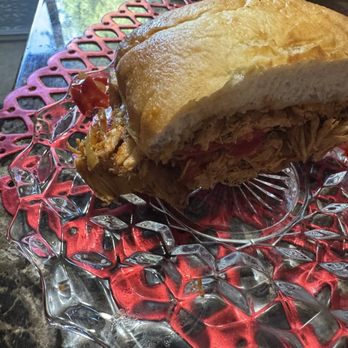 Hideout Saloon - Mothers half! - A sandwich filled with shredded pulled pork and possibly some sauce is placed on a clear glass plate. The plate rests on a red decorative placemat with a floral or geometric pattern. The sandwich bread appears toasted and the filling is visible from the side. The setting suggests a casual dining environment, possibly at a restaurant or cafe. - Cocktail Bars Near Me - Mariposa, California