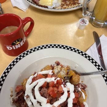 Ze's Diner - Tater tot nachos on my side of the table. Looks like a Benedict across from me. They refilled our coffee multiple times, it was great - The image shows a breakfast dish on a white plate with a black and white checkered rim, containing hash with diced tomatoes, sour cream, bacon, and fried potatoes. A fork rests on the plate. Nearby, there is a red mug labeled 'Ze's Diner' filled with coffee, and a white napkin with a knife. The setting appears to be a diner table with a light-colored surface. - Diners Near Me - Eagan, Minnesota