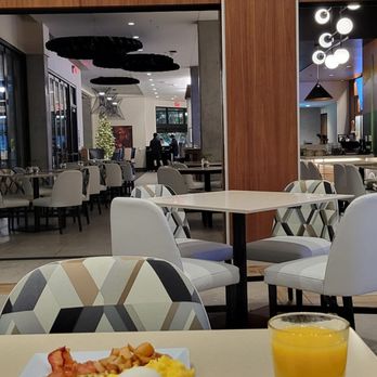 Hyatt Place Phoenix - The image shows a hotel dining area with modern furniture, including white and patterned chairs around tables. A mural wall with cacti and mountain scenery decorates one side of the space. There is a view into an adjacent lounge area with additional seating and a Christmas tree. The space is well-lit with contemporary lighting fixtures and has a clean, stylish design. - Venues & Event Spaces Near Me - Phoenix, Arizona