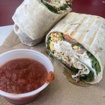 Fillmore Coffee - Breakfast Burrito - Two halves of a breakfast burrito filled with scrambled eggs, leafy greens, and meat are placed on a brown paper napkin. A small cup of red salsa is positioned nearby. The burrito is wrapped in a flour tortilla with visible grill marks, and the filling includes eggs, greens, and meat. The setting appears to be casual, likely in a cafe or restaurant. - Coffee & Tea Near Me - Phoenix, Arizona