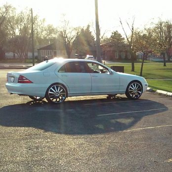 Custom Wheels - A white sedan with aftermarket wheels is parked in an empty parking lot, casting a shadow on the asphalt with a grassy area and trees in the background. - Wheel & Rim Repair Near Me - Matteson, Illinois