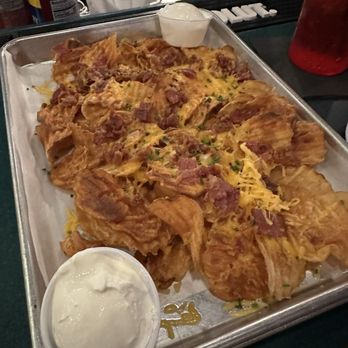 The MacStack Tavern - Loaded Potato Chips - Loaded potato chips are presented on a metal tray, topped with melted cheese, bacon bits, and chopped chives. A small cup of dipping sauce is placed on the tray. The background shows a bar or restaurant setting with various items on the counter, including a red drink container and condiments. - American Near Me - Port Clinton, Ohio