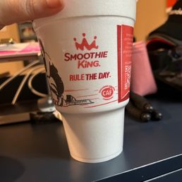a hand holding a cup of smoothie king - The image shows a white disposable cup with a red Smoothie King logo and the phrase 'RULE THE DAY' printed on it. The cup has a clear plastic lid with a straw and is held by a person's hand. The background includes a black surface, some pink papers, and various objects, indicating an indoor setting, possibly an office or workspace.