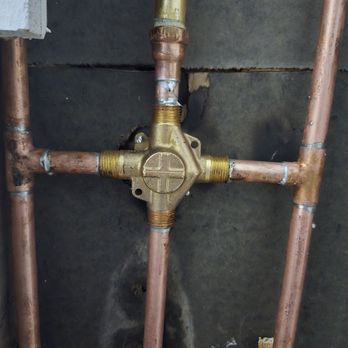 Superior Rooter - New shower valve - Copper pipes are arranged in a cross configuration, connected to a brass valve in the center. The pipes and valve are mounted on a dark, textured wall, with visible signs of oxidation, corrosion, and wear on the joints. The setup appears to be part of a plumbing system, possibly in a building's interior wall. - Plumbing Near Me