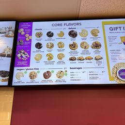 Digital menu board at Insomnia Cookies features cookie flavors categorized as classic, deluxe, vegan, and gluten-free, with images and calorie information. Beverages such as soda, water, milk, and Powerade are listed with prices. The menu includes options like chocolate chunk, snickerdoodle, s'mores deluxe, and vegan chocolate chunk, among others. The display is mounted on a wall with a purple and white color scheme, and part of the surrounding wall is visible.