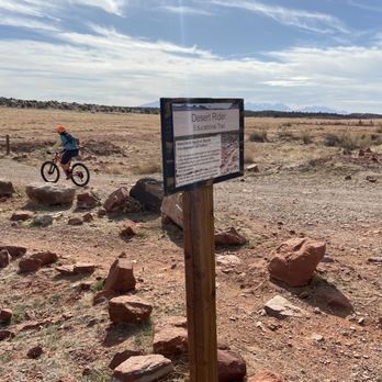 Moab Cliffs & Canyons - A cyclist wearing a helmet and backpack rides a mountain bike on a dirt trail in a desert environment. The trail is rocky with scattered stones and dry vegetation. A sign in the foreground reads 'Desert Rider Educational Trail,' providing information about the area. The background features an expansive desert landscape with distant mountains under a partly cloudy sky, indicating an outdoor recreational area focused on mountain biking and desert exploration. - Climbing Near Me - Moab, Utah