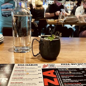 Crust Brewing - A clear glass water bottle and a black mug with a straw and mint garnish are placed on a wooden restaurant table. The menu in front of the items lists various food options. In the background, there are patrons seated at the bar, some wearing hats, and multiple television screens displaying sports. The setting appears lively and casual, with a focus on food and drinks. - Breweries Near Me - Rosemont, Illinois