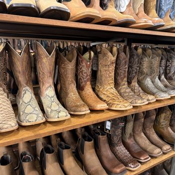 Boot Barn - Exotic western boots - A retail display features multiple pairs of cowboy boots arranged on wooden shelves. The boots showcase various western styles, including snakeskin patterns, embossed leather, and traditional leather finishes. The display includes boots with pointed toes, decorative stitching, and different shades of brown, tan, and beige. The boots are organized in rows, with some pairs on the upper shelf and others on the lower shelf, highlighting a variety of western footwear options. - Shoe Stores Near Me - Cincinnati, Ohio