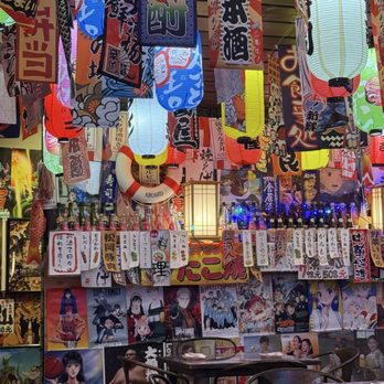 Dragon Sushi - Indoor seating - The image shows the interior of a Japanese restaurant decorated with numerous colorful paper lanterns and banners featuring Japanese characters. The walls are adorned with anime posters and artwork, creating a vibrant atmosphere. There are tables and chairs in the foreground, with some bottles and a life preserver hanging on the wall. The lighting is warm, and the overall decor emphasizes Japanese cultural themes. - Sushi Bars Near Me - Albuquerque, New Mexico