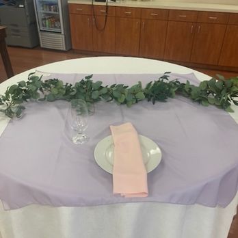 HEB - My color scheme - A round table is covered with a white tablecloth and a purple table runner. It is set with a white plate, a pink napkin, and a clear glass. A greenery garland runs across the center of the table as decoration. In the background, there is a wooden counter with various items, including a phone, flowers, and bottles, and a beverage refrigerator is visible to the left. - Grocery Near Me - Pflugerville, Texas