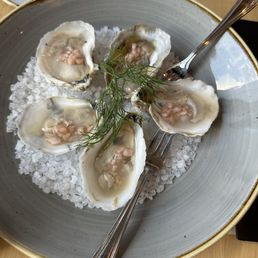Oysters - A dish of five raw oysters on a bed of crushed ice, garnished with a sprig of herbs, served in a large bowl with a fork and spoon.
