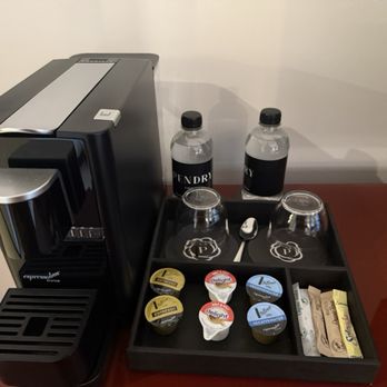 Pendry Chicago - A coffee station includes a black espresso machine, two bottled waters labeled Pendry, and a tray with two upside-down cups, a spoon, and assorted coffee pods and creamers. The setup is on a wooden surface against a plain wall. - Hotels Near Me - Chicago, Illinois