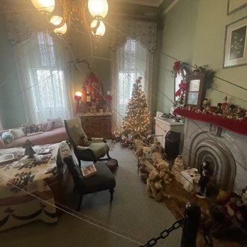 David Davis  Mansion - The image shows a living room decorated for Christmas, with a small, lit Christmas tree in the corner. The room includes a sofa with plush toys and pillows, a table with holiday-themed items, and chairs. The fireplace mantel is adorned with figurines and a red cloth, and there are holiday decorations on a dresser and wall. The room has lace curtains, a chandelier, and framed pictures on the walls. - Museums Near Me - Bloomington, Illinois