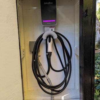 Infinite Electric - An electric vehicle charging station, JuiceBox model, is installed inside a cabinet with a white interior. The charger has a black exterior with purple indicator lights and a coiled charging cable hanging from it. The cabinet appears to be outdoors, with some greenery visible on the right side of the image. - Electricians Near Me - Manassas, Virginia