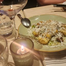 A plate of gnocchi topped with shredded cheese, herbs, and possibly olive oil is placed on a wooden dining table. The table includes a lit candle in a glass holder, a glass of red wine, a water glass, and other plates with remnants of food. The setting suggests a casual dining environment with multiple dishes and drinks.