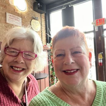 Prost - Two women are smiling and posing for a selfie inside a restaurant or cafe. The woman on the left has white hair, wears glasses with pink frames, and is dressed in a red sweater. The woman on the right has short, reddish hair and is wearing a light green sweater. Behind them, there is a Christmas tree decorated with ornaments, and the interior features brick walls, large windows, and various signs, including an exit sign and a ticket office sign. - Bars Near Me - Bellevue, Kentucky