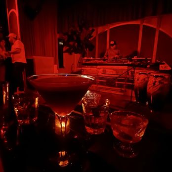 Hoy Como Ayer - A dark bar scene featuring cocktails on the table and a DJ playing music in the background. - Music Venues Near Me - Miami, Florida