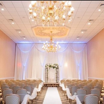 Meridian Banquets - Ceremony room - A spacious wedding ceremony room with high ceilings, ornate chandeliers, and soft ambient lighting. The back wall is decorated with white draped curtains and a floral arch. Rows of chairs with patterned upholstery are arranged facing the altar, with white ribbons tied to the chairs. The room has a neutral color palette with beige walls and a patterned carpet, creating an elegant setting for a wedding. - Venues & Event Spaces Near Me - Rolling Meadows, Illinois