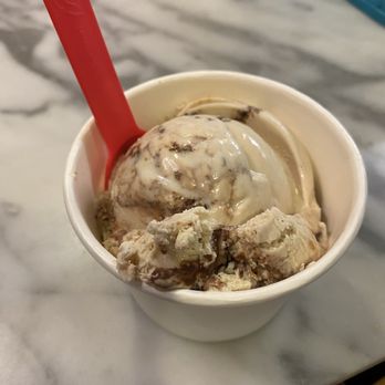 Churn - I can't remember the flavor but it was yummy - A small white cup contains a scoop of ice cream with visible chocolate swirls and chunks. A red plastic spoon is inserted into the ice cream. The cup is placed on a marble surface, with a blurred background that includes a blue object and some packaging at the bottom edge. - Ice Cream & Frozen Yogurt Near Me - Phoenix, Arizona