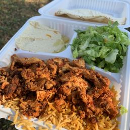 Chicken Shawarma Plate - A white disposable food container is filled with seasoned rice topped with cooked, seasoned meat, alongside a fresh green salad and flatbread. The container is open, revealing the food inside, and is held outdoors on a grassy area with sunlight illuminating the scene.