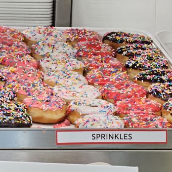 Shipley Do-Nuts - A metal tray holds numerous donuts decorated with various icing and colorful sprinkles. The donuts have pink, white, and chocolate icing, arranged in rows. The display appears to be in a bakery or donut shop setting, with a label indicating 'Sprinkles'. - Donuts Near Me - Leander, Texas