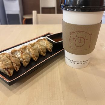 Gom Ramen - Mandu - A paper coffee cup with a black plastic lid and a cardboard sleeve featuring a bear face illustration is positioned on a light wooden table. Next to the coffee is a rectangular black tray holding several gyoza dumplings arranged in a row, accompanied by a small dish of dipping sauce. The background includes chairs and a water dispenser, indicating a casual dining or cafe setting. - Noodles Near Me - El Paso, Texas