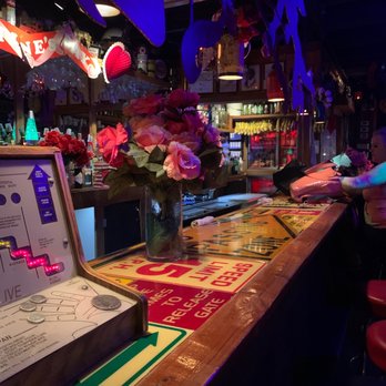 Nu Towne Saloon - a woman sitting at the bar - The image shows a bar counter with a glass vase containing pink and purple flowers. There are illuminated signs and decorations hanging above the counter, including a heart-shaped light. Behind the bar, shelves stocked with bottles and glasses are visible. A person on the right side is reaching for a pink bag, with their arm extended across the counter. The setting appears to be a lively bar environment with dim lighting and decorative elements. - Gay Bars Near Me - Phoenix, Arizona