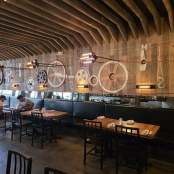 Bodegon No 69 - The image shows the interior of a restaurant with a wooden ceiling and a large mirror on the wall reflecting the space. The wall is decorated with large gear-shaped designs and industrial-style lighting fixtures. There are tables set with napkins and utensils, and a few patrons seated at one of the tables. The overall decor combines industrial and modern elements, creating a distinctive ambiance. - Latin American Near Me - Pasadena, California