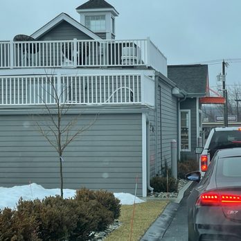 Dunkin' -  a car parked in front of a house - The image shows a gray, two-story building with horizontal siding, featuring a rooftop deck enclosed by white railings. The deck has a small tower-like structure with windows. In front of the building, there is a landscaped area with bushes, a small leafless tree, and patches of snow. Several cars are parked along the side of the building, with one car's taillights visible. The sky is overcast, and utility poles and wires are visible in the background. - Coffee & Tea Near Me - Tolland, Connecticut