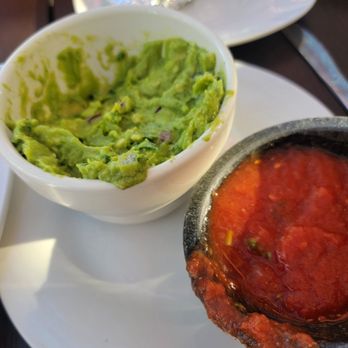 Fiesta Jalapenos - The image shows a white plate with two small dishes of Mexican condiments: guacamole in a white bowl and salsa in a black stone dish. The guacamole appears freshly made with a chunky texture, while the salsa is red with visible pieces of tomato and chili. In the background, there is a foil-wrapped item and a white plate, suggesting a meal setting at a Mexican restaurant. - Mexican Near Me - Ankeny, Iowa