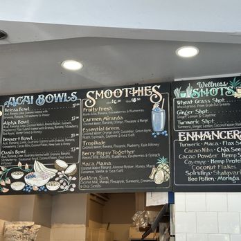 Main Squeeze - Menu May 2025 - A black chalkboard-style menu board features sections for juices, smoothies, wellness shots, and enhancers at a cafe near Lake Merritt, Oakland, CA. The menu includes items like acai bowls, fruit smoothies, and wellness shots such as wheatgrass and ginger shots, with detailed ingredients and prices. The board is mounted on a wall with a white ceiling above, illuminated by ceiling lights, and has decorative illustrations of ingredients and drinks. - Juice Bars & Smoothies Near Me - Oakland, California