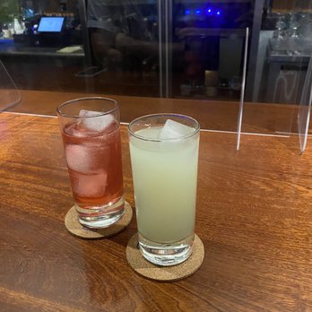The Watershed - Pale Stranger and Cherry Hibiscus Sparkler - Two cocktails with ice are placed on cork coasters on a wooden bar counter, separated by a clear acrylic divider, in a bar setting. - Bars Near Me - Ithaca, New York