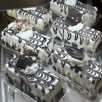 Bilqis Coffee - Divine pastries - Several slices of layered cake with white icing and chocolate drizzle are displayed in clear plastic containers. Each slice is topped with whipped cream and chocolate decorations, including chocolate squares and swirls. The cakes are arranged on a reflective surface, likely in a bakery or dessert display case. - Coffee & Tea Near Me - Brooklyn, New York