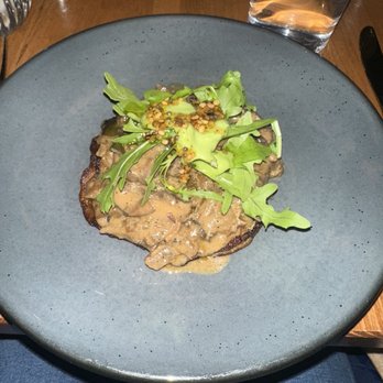 Lore - Mushroom dish - A mushroom dish with a creamy sauce is presented on a dark ceramic plate. It is topped with fresh arugula and other greens, and sprinkled with mustard seeds. The setting includes a glass of water and a wooden table, indicating a restaurant environment. - Indian Near Me - Brooklyn, New York