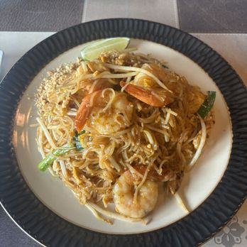 Little Thai Corner - Super Pad Thai - A plate of Pad Thai featuring shrimp, bean sprouts, chopped peanuts, and green onions on a black-rimmed dish. The dish appears to be stir-fried with a glossy sauce, and a lime wedge is placed on the side for added flavor. The plate is set on a table with a patterned tablecloth, and some utensils and a partially eaten dish are visible in the background. - Thai Near Me - Warwick, Rhode Island