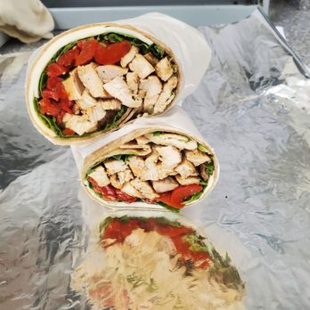 Sabeli - Two chicken wraps filled with grilled chicken, tomatoes, and greens are shown on aluminum foil, with a reflective surface underneath. - Delis Near Me - Syosset, New York