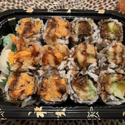 a tray of sushi and a glass of beer - A black plastic take-out container features an assortment of sushi rolls with various fillings, including vegetables and possibly cooked seafood. The rolls are topped with sauces, and some have visible cucumber and other ingredients. The container is placed on a textured surface, and there are garnishes such as pickled ginger and wasabi on the side.