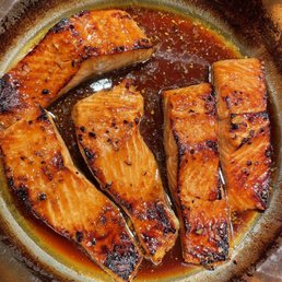 King Salmon - Four pieces of salmon fillet are shown in a pan, cooked with a dark, glossy sauce that appears to be a glaze or marinade. The salmon has a caramelized exterior with some charring, indicating it was seared or broiled. The sauce surrounds the fish, with some oil or juices visible, suggesting a flavorful preparation.